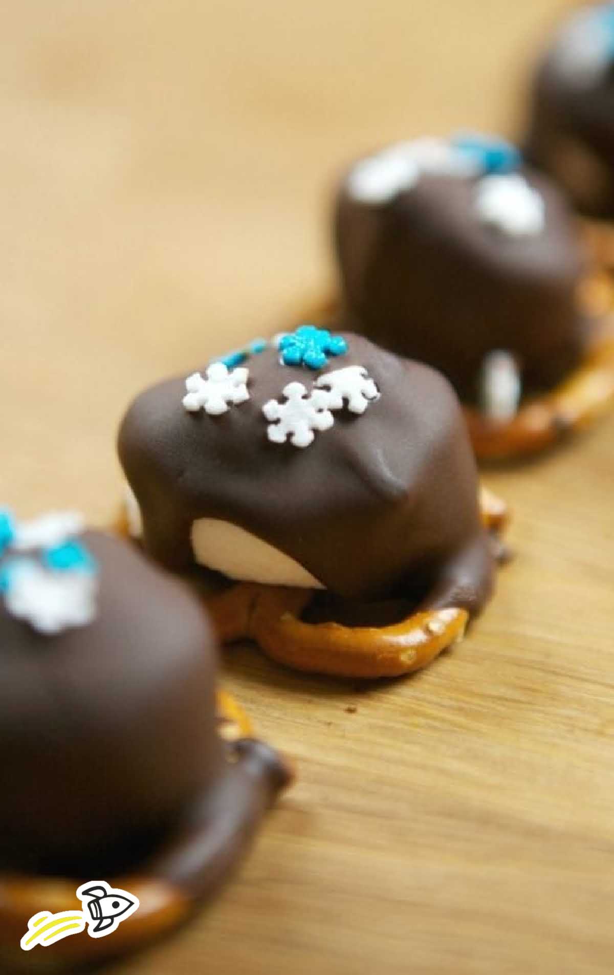 Row of chocolate-covered marshmallows with sprinkles on top of mini pretzels, displayed on a wooden surface.