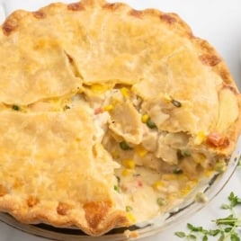 Whole chicken pot pie with a slice removed, showing creamy chicken and vegetable filling with a side of fresh thyme
