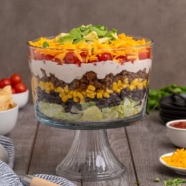 A trifle dish filled with Mexican Layered Salad, featuring lettuce, black beans, corn, seasoned ground beef, tomatoes, creamy sauce, shredded cheese, and avocado, set on a wooden table with toppings and bowls nearby.