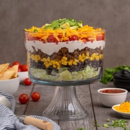 A glass trifle bowl filled with Mexican Layered Salad—lettuce, black beans, corn, seasoned ground beef, sour cream, diced tomatoes, and shredded cheese—garnished with cilantro sits on a wooden table.