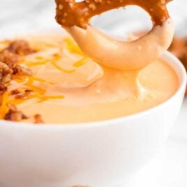 close up shot of a pretzels dipped into a condiment bowl of Beer cheese dip topped with shredded cheese and bacon bits