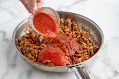 A hand pours tomato sauce from a measuring cup into a stainless steel skillet filled with browned ground meat, the perfect start for classic Baked Ziti, all set on a white marble countertop.