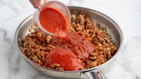 A hand pours tomato sauce from a measuring cup into a stainless steel skillet filled with browned ground meat, the perfect start for classic Baked Ziti, all set on a white marble countertop.