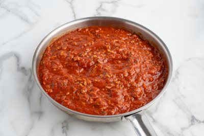 A stainless steel skillet filled with chunky red tomato sauce, perfect for Baked Ziti, sits on a white marble countertop. The sauce appears to contain herbs and pieces of tomato.