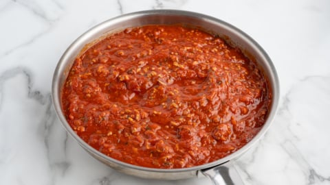 A stainless steel skillet filled with chunky red tomato sauce, perfect for Baked Ziti, sits on a white marble countertop. The sauce appears to contain herbs and pieces of tomato.