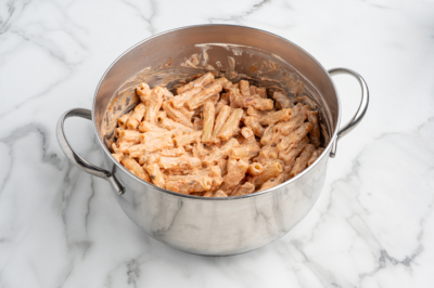 A stainless steel pot filled with creamy pasta, likely baked ziti or rigatoni, coated in a light orange or pink sauce, sits on a white marble surface.