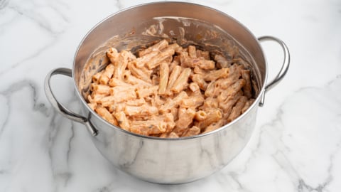 A stainless steel pot filled with creamy pasta, likely baked ziti or rigatoni, coated in a light orange or pink sauce, sits on a white marble surface.