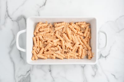 A white rectangular baking dish filled with uncooked pasta coated in a creamy orange sauce, ready for the oven—perfect for making classic Baked Ziti—sits on a white marble surface.