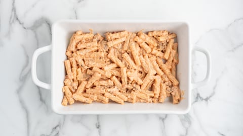 A white rectangular baking dish filled with uncooked pasta coated in a creamy orange sauce, ready for the oven—perfect for making classic Baked Ziti—sits on a white marble surface.