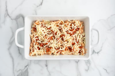 A white rectangular baking dish on a marble surface, filled with shredded cheese and pieces of chicken mixed with tomato sauce, perfect for preparing a delicious Baked Ziti.