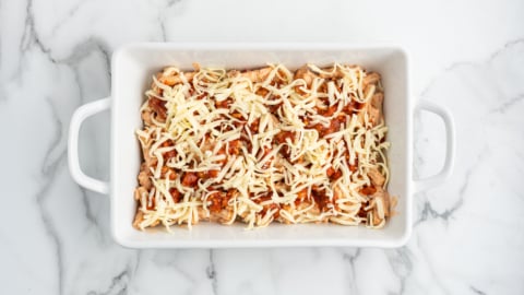 A white rectangular baking dish on a marble surface, filled with shredded cheese and pieces of chicken mixed with tomato sauce, perfect for preparing a delicious Baked Ziti.