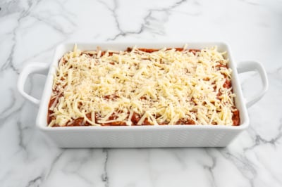 A white baking dish filled with unbaked lasagna, topped with shredded cheese, sits on a marble countertop, ready to be baked like a classic Baked Ziti.