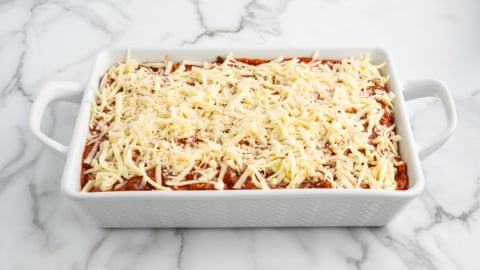 A white baking dish filled with unbaked lasagna, topped with shredded cheese, sits on a marble countertop, ready to be baked like a classic Baked Ziti.
