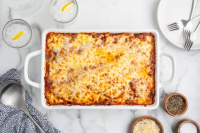 A baked lasagna in a white rectangular dish on a marble table, surrounded by a serving spoon, napkin, plates, forks, glasses with ice and lemon, and small bowls of cheese and pepper—perfect for fans of baked ziti.