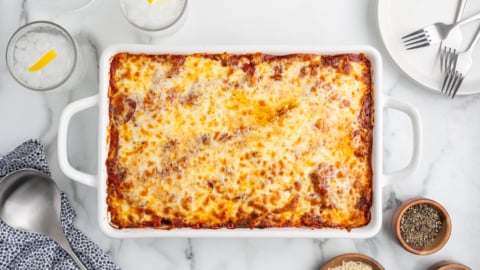 A baked lasagna in a white rectangular dish on a marble table, surrounded by a serving spoon, napkin, plates, forks, glasses with ice and lemon, and small bowls of cheese and pepper—perfect for fans of baked ziti.