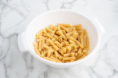 Uncooked rigatoni pasta in a white plastic colander sits on a white marble countertop, ready to be used in a classic Baked Ziti recipe.