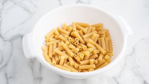 Uncooked rigatoni pasta in a white plastic colander sits on a white marble countertop, ready to be used in a classic Baked Ziti recipe.