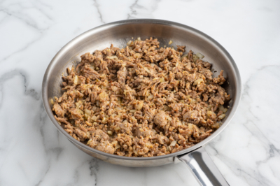 Cooked ground beef mixed with chopped onions in a stainless steel skillet, perfect as a hearty base for Baked Ziti, placed on a white marble countertop.