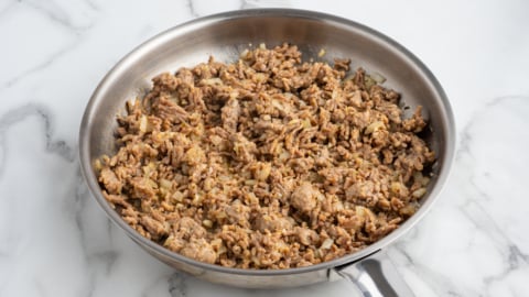 Cooked ground beef mixed with chopped onions in a stainless steel skillet, perfect as a hearty base for Baked Ziti, placed on a white marble countertop.