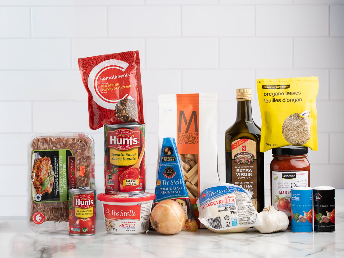 Various Italian cooking ingredients for Baked Ziti, including ground beef, ricotta, mozzarella, dried pasta, tomato sauce, olive oil, an onion, garlic, dried oregano, marinara sauce, and canned tomatoes on a white background.