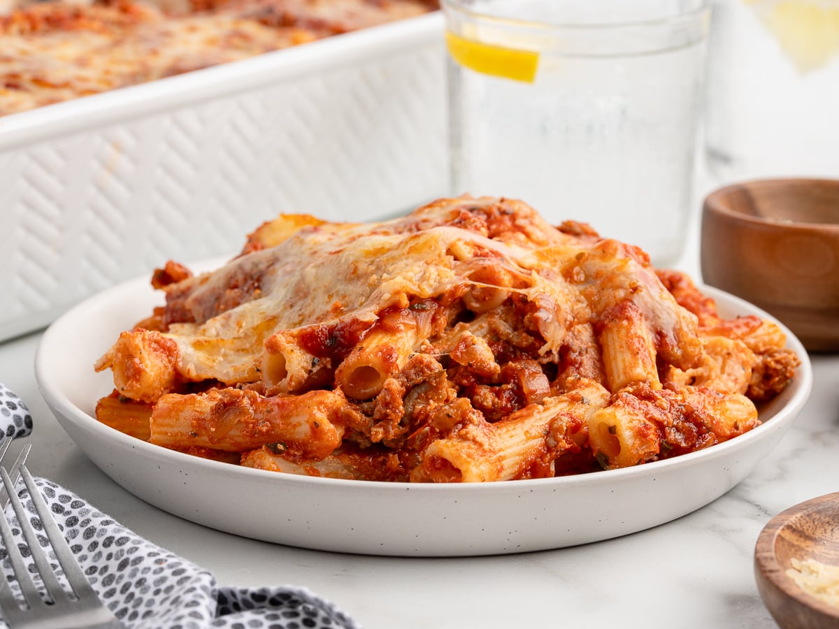 A plate of Baked Ziti pasta with melted cheese and tomato sauce sits on a white table near a glass of water with lemon, with a bubbling Baked Ziti baking dish in the background.