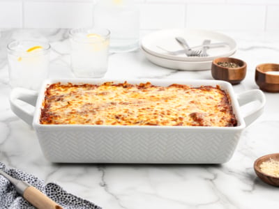 A baked lasagna with a golden, melted cheese topping sits in a white rectangular dish on a marble counter, surrounded by plates, utensils, drinks, and small bowls of seasoning—perfect for fans of classic Baked Ziti.