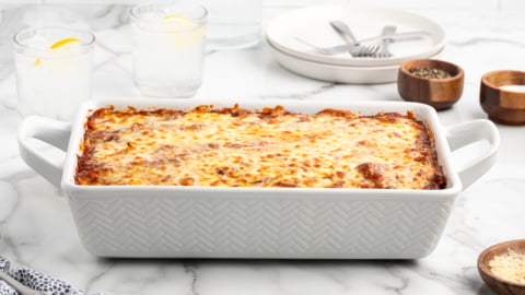 A baked lasagna with a golden, melted cheese topping sits in a white rectangular dish on a marble counter, surrounded by plates, utensils, drinks, and small bowls of seasoning—perfect for fans of classic Baked Ziti.