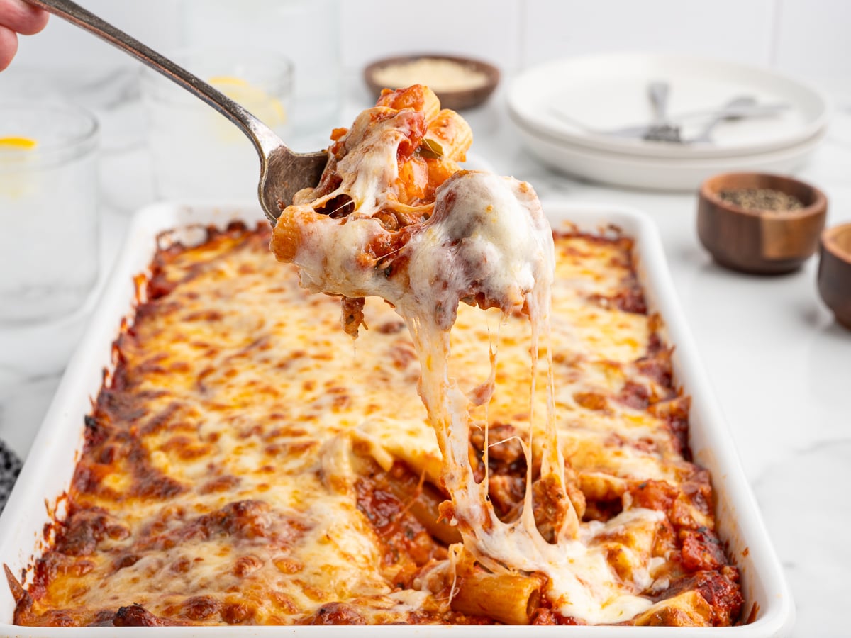 A close-up of cheesy Baked Ziti being served from a white baking dish. Melted cheese stretches from the spoon, revealing ziti pasta in tomato sauce beneath a golden, bubbly cheese topping. Plates and seasoning bowls are in the background.