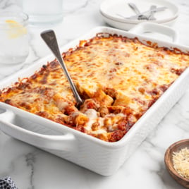 A white baking dish filled with cheesy baked ziti sits on a marble surface, with a metal serving spoon lifting a portion. Plates, glasses of water, grated cheese, and pepper are arranged nearby.
