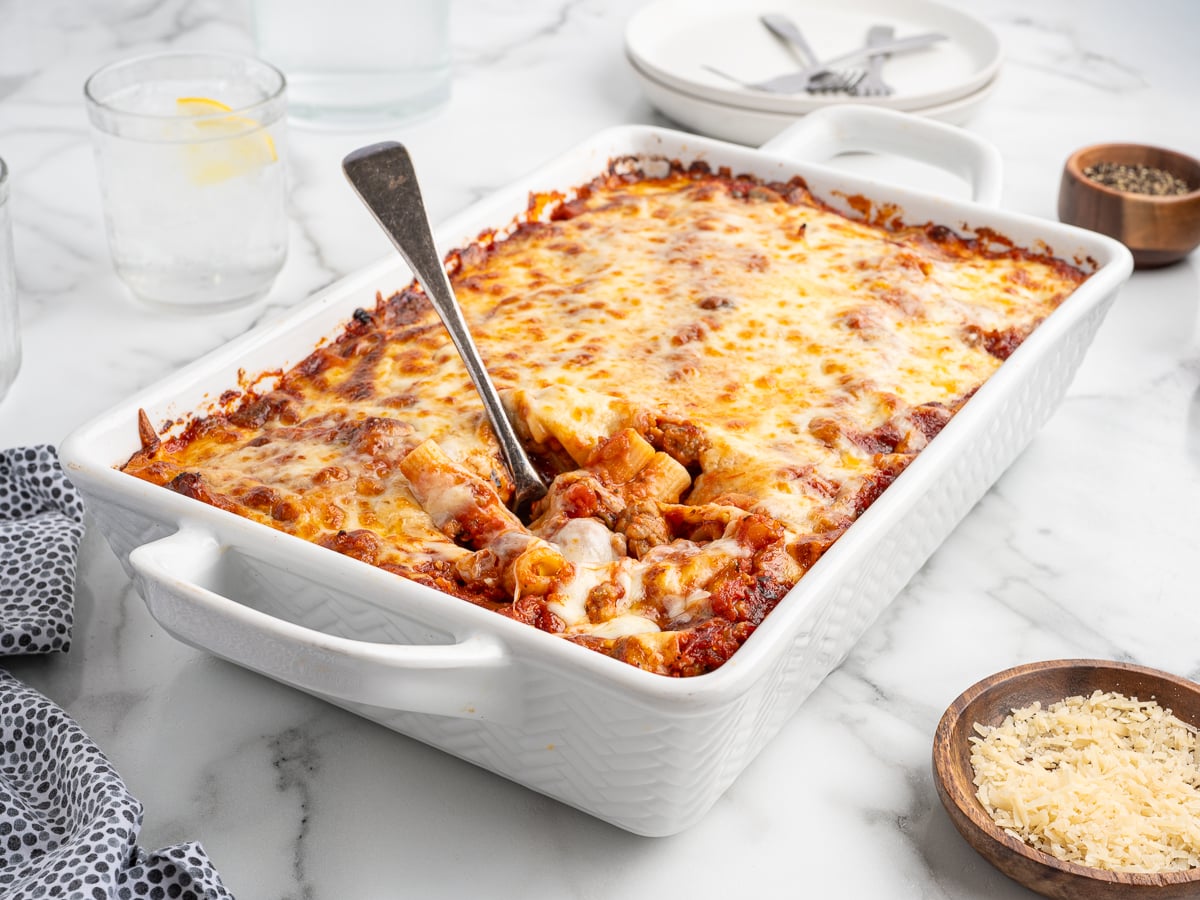 A white baking dish filled with cheesy baked ziti sits on a marble surface, with a metal serving spoon lifting a portion. Plates, glasses of water, grated cheese, and pepper are arranged nearby.