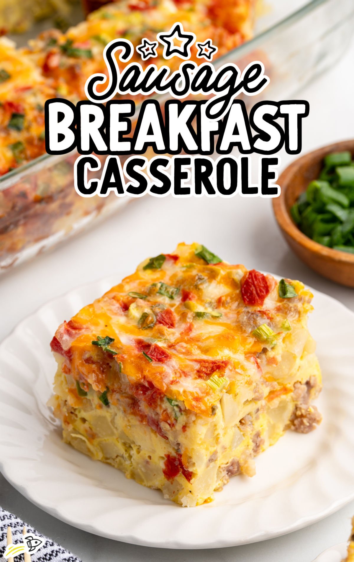 A square slice of sausage breakfast casserole on a white plate, showing layers of eggs, potatoes, sausage, melted cheese, and diced red and green peppers.