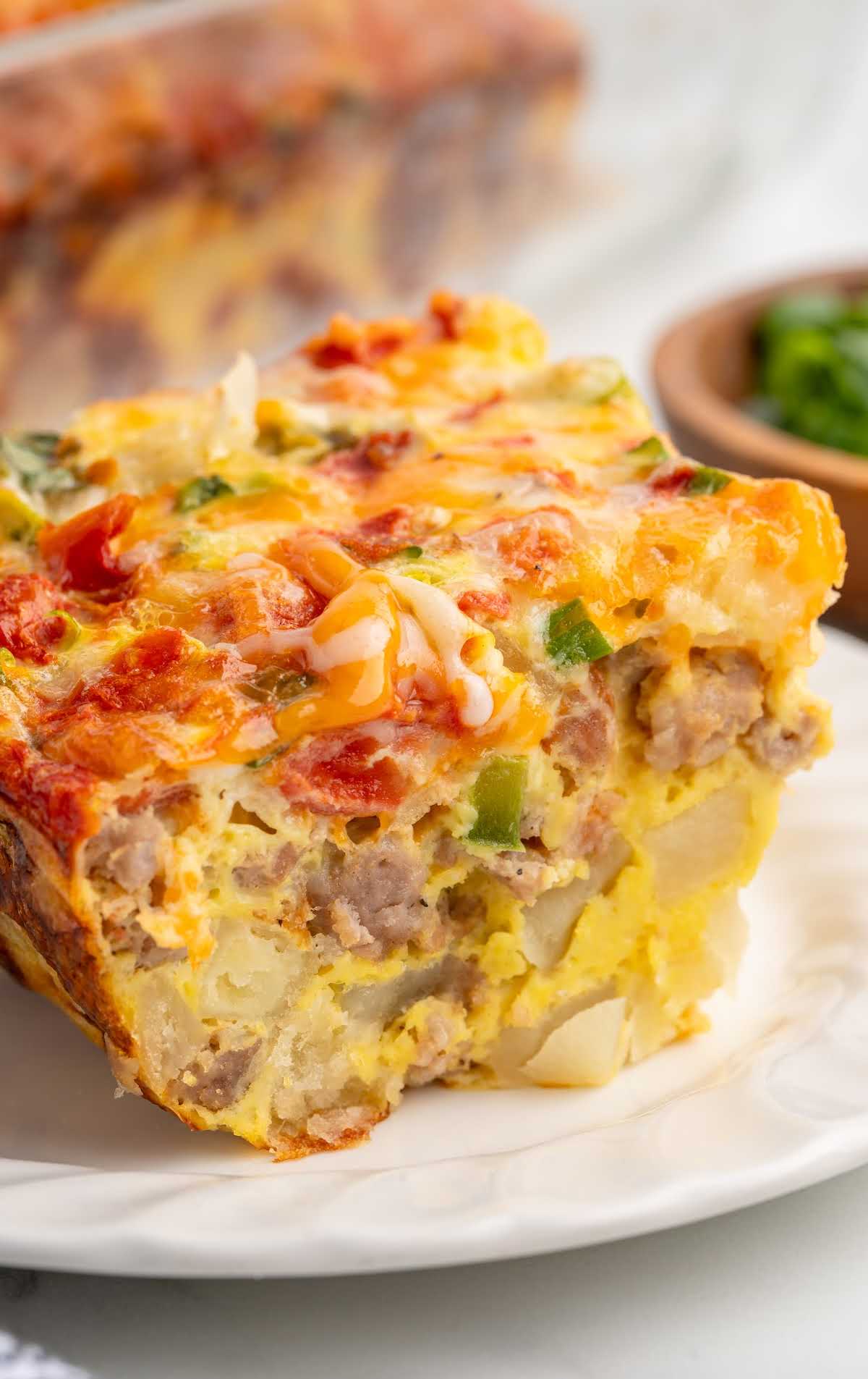 Close up of sausage breakfast casserole slice on a white plate with fork, filled with eggs, sausage, and vegetables.