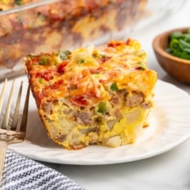 Slice of sausage breakfast casserole on a white plate with fork, showing layers of eggs, sausage, potatoes, and melted cheese.