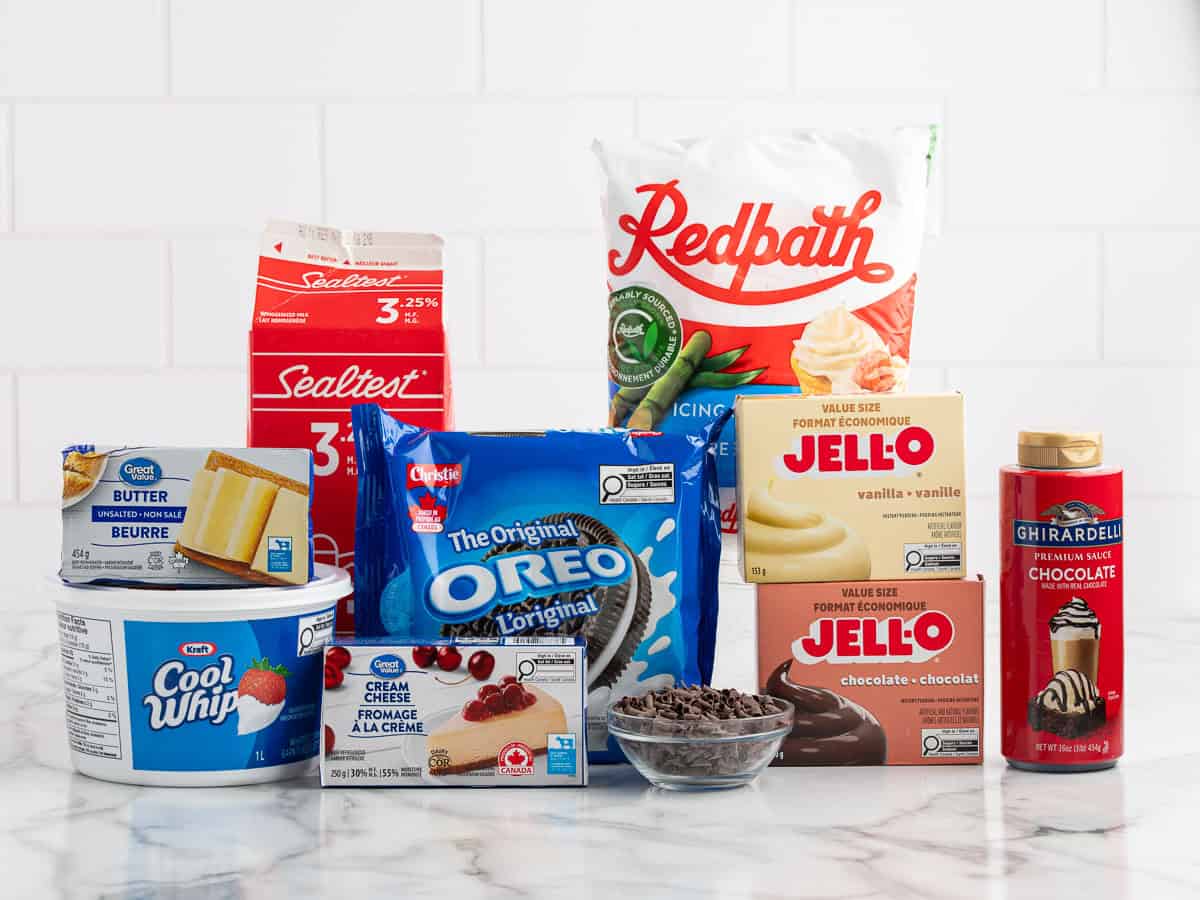 Assorted dessert ingredients on a counter, including Cool Whip, cream cheese, butter, chocolate chips, Oreos, Jello puddings, milk, granulated sugar, and chocolate syrup against a white tile background—everything you need for the ultimate Chocolate Lasagna Recipe.