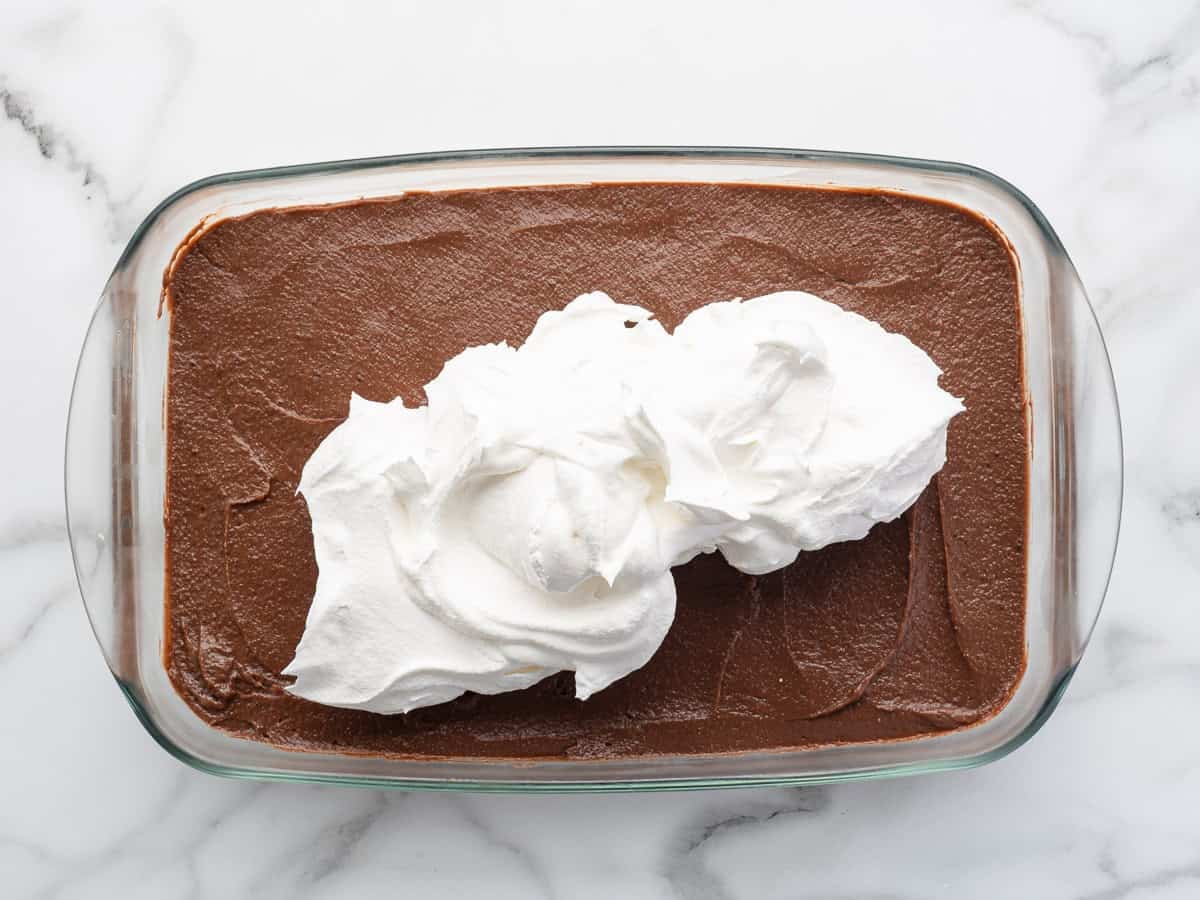 A glass dish filled with chocolate pudding is topped with a large dollop of whipped cream, creating a simple twist reminiscent of a classic Chocolate Lasagna. All placed on a white marble surface, this dessert brings the comforting flavors of your favorite Chocolate Lasagna Recipe to life.