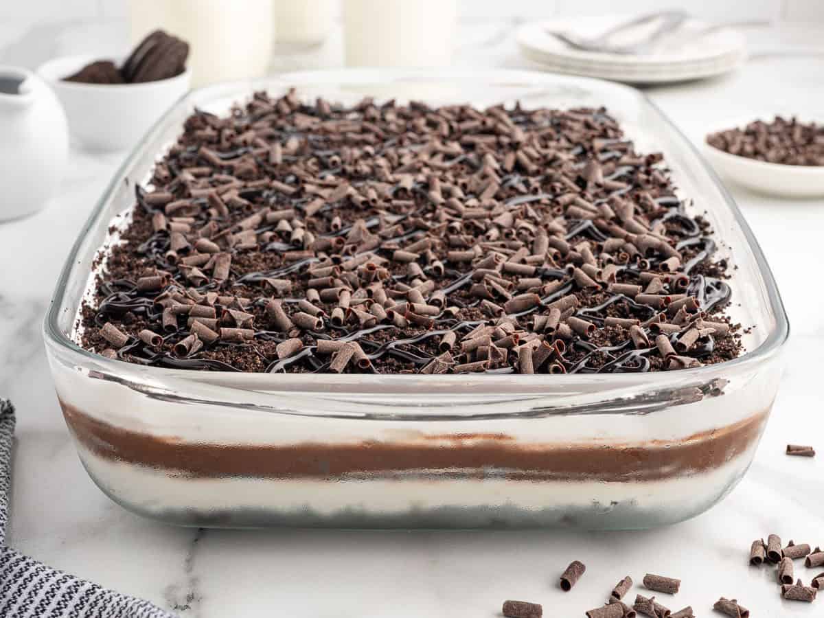 A glass dish filled with layered chocolate dessert, reminiscent of a classic Chocolate Lasagna Recipe, is topped with chocolate curls and shavings. Plates and bowls with extra chocolate can be seen in the background.