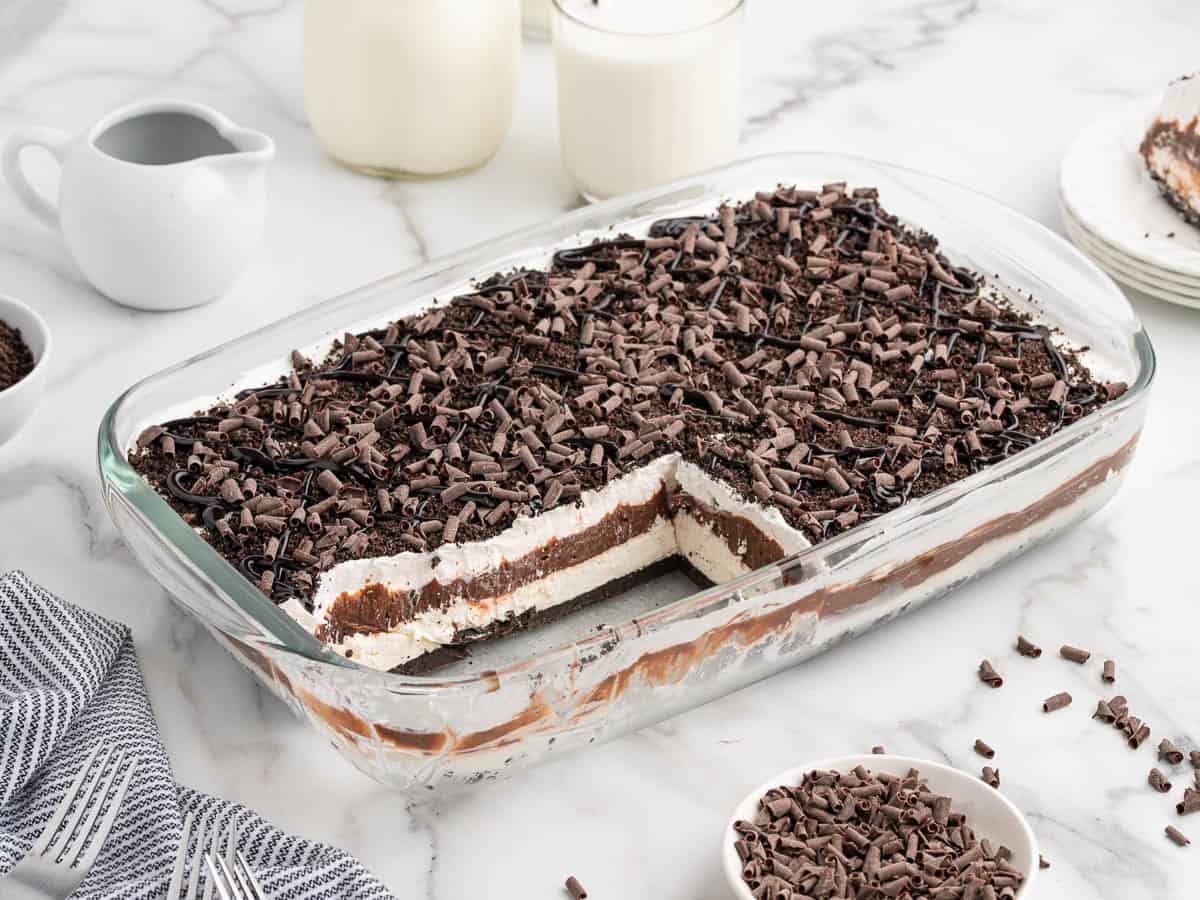 A glass dish of layered chocolate dessert, reminiscent of a decadent Chocolate Lasagna recipe, with a section removed to reveal layers of cream, pudding, and chocolate curls on top. A bowl of chocolate curls and glasses of milk are nearby on a marble countertop.