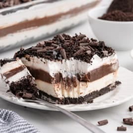 A decadent Chocolate Lasagna sits on a white plate, showcasing its luscious layers of chocolate, cream, and cookie crust. A fork has taken a bite from the dessert, revealing its rich texture beneath a sprinkle of chocolate curls. In the background, bowls filled with cookies and extra chocolate curls offer hints of the irresistible Chocolate Lasagna Recipe used to create this indulgent treat.