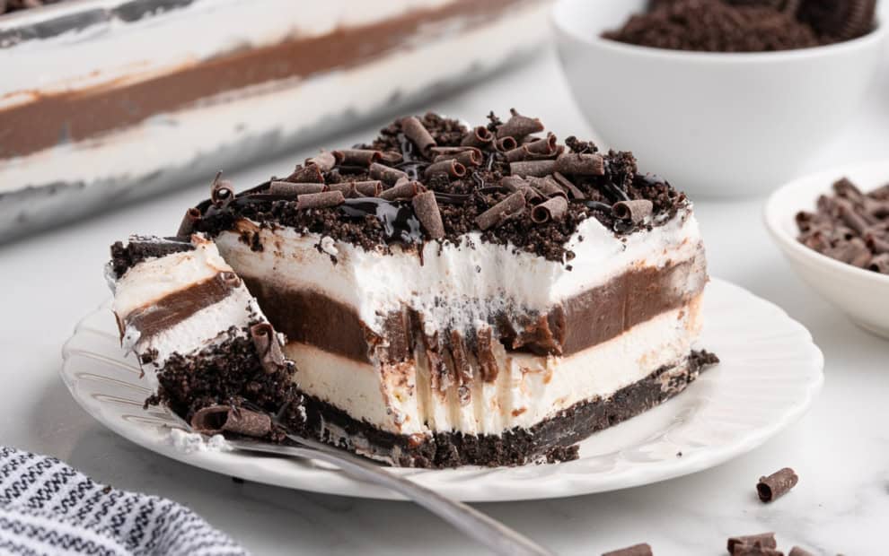 A decadent Chocolate Lasagna sits on a white plate, showcasing its luscious layers of chocolate, cream, and cookie crust. A fork has taken a bite from the dessert, revealing its rich texture beneath a sprinkle of chocolate curls. In the background, bowls filled with cookies and extra chocolate curls offer hints of the irresistible Chocolate Lasagna Recipe used to create this indulgent treat.