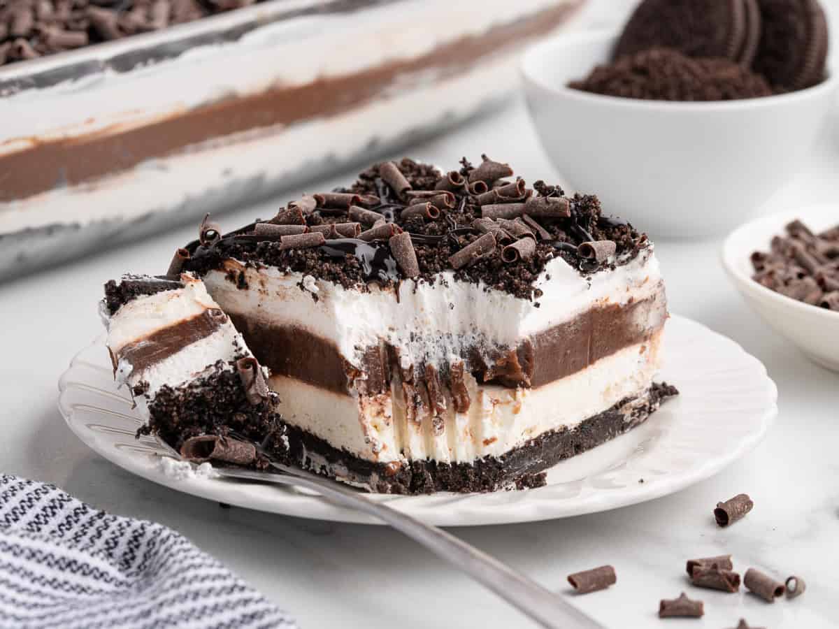A decadent Chocolate Lasagna sits on a white plate, showcasing its luscious layers of chocolate, cream, and cookie crust. A fork has taken a bite from the dessert, revealing its rich texture beneath a sprinkle of chocolate curls. In the background, bowls filled with cookies and extra chocolate curls offer hints of the irresistible Chocolate Lasagna Recipe used to create this indulgent treat.