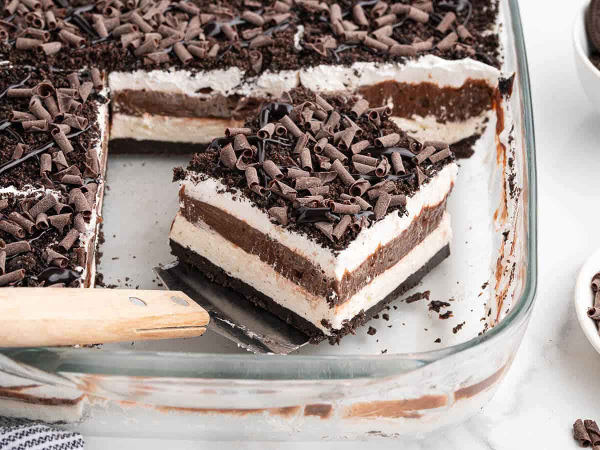 A glass dish with layered Chocolate Lasagna dessert topped with whipped cream, chocolate shavings, and cookie crumbs. A square slice is lifted out with a spatula, showcasing the irresistible layers in this classic Chocolate Lasagna Recipe.