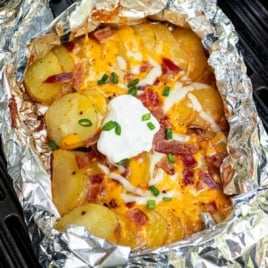 Overhead view of sliced Yukon gold potatoes, shredded cheeses, bacon, and seasonings arranged on a wooden cutting board for campfire potatoes recipe.