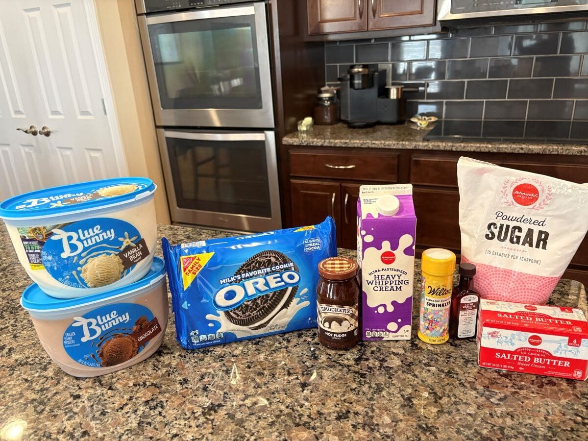 Ingredients for a Copycat Dairy Queen Ice Cream Cake&mdash;Oreos, chocolate syrup, ice cream, heavy whipping cream, vanilla extract, powdered sugar, butter, and nonstick spray&mdash;are set out on a granite kitchen counter with stove and cabinets in the background.