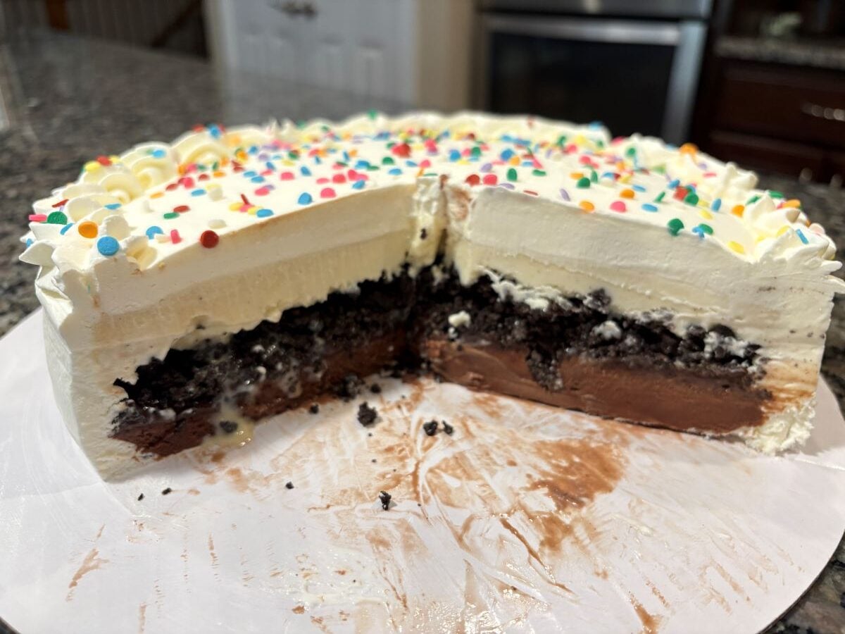 A partially sliced Copycat Dairy Queen Ice Cream Cake with layers of chocolate and vanilla, topped with whipped cream and colorful sprinkles, sits on a white surface in a kitchen.