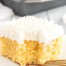 close up shot of coconut poke cake with coconut frosting on a white plate with a piece taken out of it with a fork