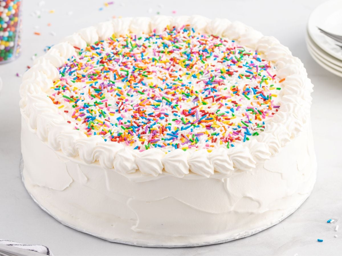 A round vanilla cake inspired by a Copycat Dairy Queen Ice Cream Cake, covered in white frosting and topped with colorful rainbow sprinkles. Piped decorative edges complete the look, with some plates visible in the background.