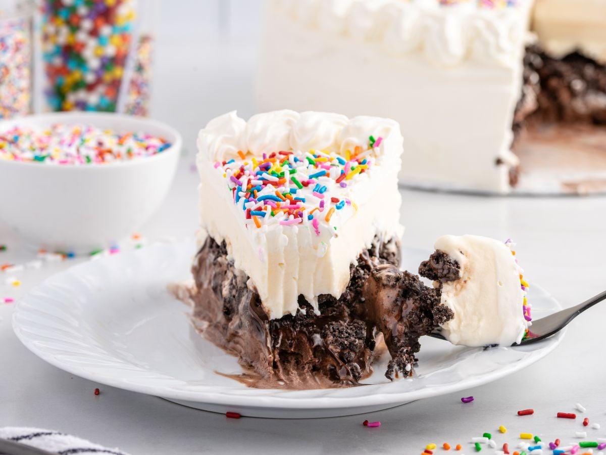 A slice of Copycat Dairy Queen Ice Cream Cake with chocolate and vanilla layers, topped with whipped cream and colorful sprinkles, sits on a white plate. A fork holds a bite of cake, while a bowl of sprinkles is in the background.