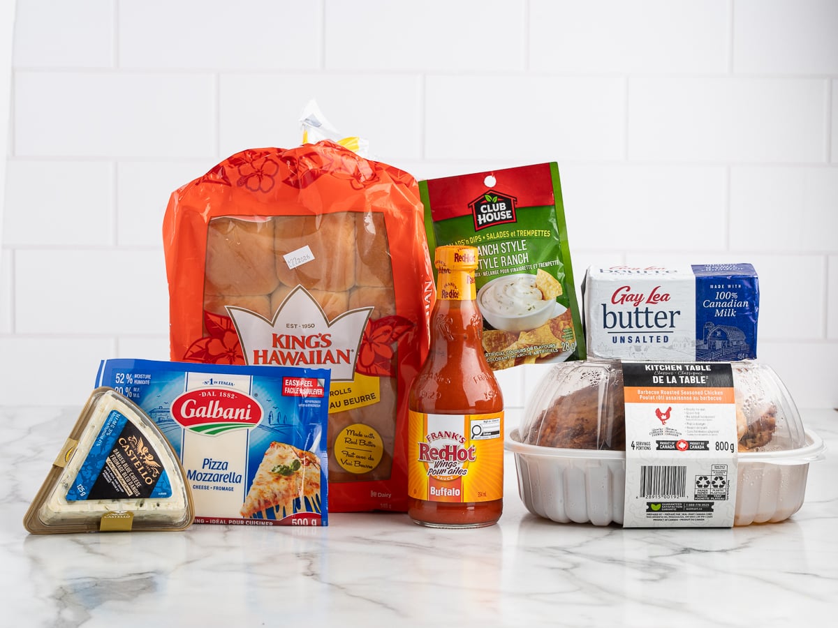 A variety of groceries including Kings Hawaiian rolls, Franks RedHot Buffalo sauce&mdash;perfect for a Buffalo Chicken Sliders recipe&mdash;Gay Lea butter, rotisserie chicken, Club House gravy mix, Galbani pizza mozzarella, blue cheese, and kitchen table cookies.