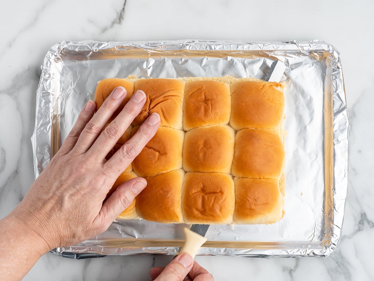 A hand holds a group of dinner rolls on a foil-lined baking sheet while another uses a spatula to separate the rolls, perfect for assembling your Buffalo Chicken Sliders.