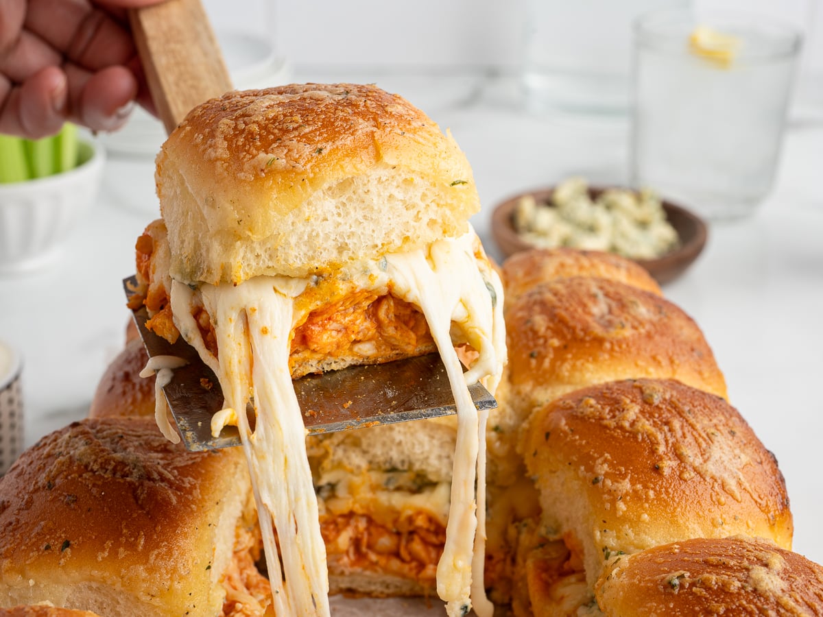 A hand lifts a cheesy, golden-brown Buffalo Chicken Slider filled with melted cheese and shredded chicken from a round baking dish, with gooey cheese stretching between the buns&mdash;an irresistible Buffalo Chicken Sliders recipe in action.